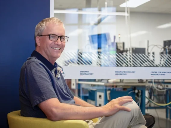 Dr. Michael Lesser smiling and sitting in a chair with a window to the ITL clean room in the background at the Applied Research Building.