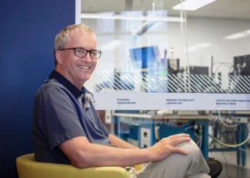 Dr. Michael Lesser smiling and sitting in a chair with a window to the ITL clean room in the background at the Applied Research Building.