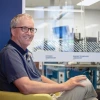 Dr. Michael Lesser smiling and sitting in a chair with a window to the ITL clean room in the background at the Applied Research Building.